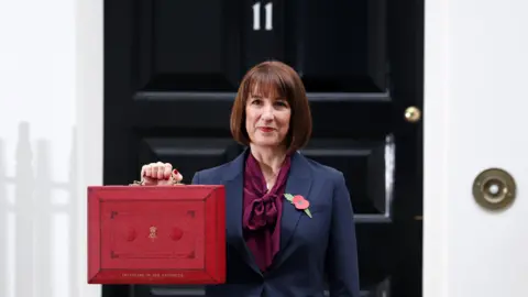 EPA-EFE/REX/Shutterstock Chancellor Rachel Reeves holds up her red budget box outside the black door to number 11 Downing Street on Wednesday morning, before heading to the House of Commons to make her statement. The box has the words "Chancellor of the Exchequer" printed on it in gold lettering.