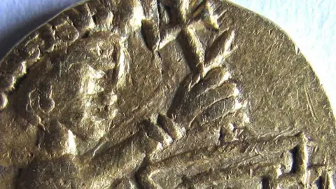 A close up of a 7th Century gold coin. It shows a male figure with an oversized head, wearing a Roman-style headdress or diadem. Its left arm is raised up and appears to be holding a cross. 