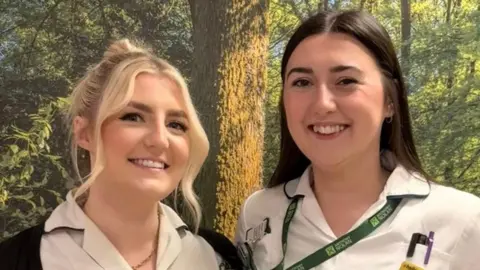 CUH Two women standing next to each other in front of a wall which has woodland themed wallpaper. The woman on the left has blonde hair in a ponytail and is wearing a white healthcare T-shirt and a black cardigan. The woman on the right has long brown hair and is wearing a white healthcare T-shirt and a green lanyard.