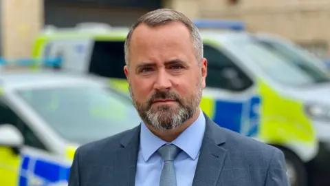 Portrait photograph of DCI Neil Third standing by police cars