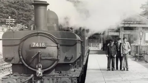 A black and white image of a steam train waiting on a platform while three men have their arms round each other smiling for a photo. They are wearing suits and hats, that look like they work at the station.