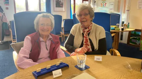 An elderly woman wearing a pink jumper and red body warmer sits at at a table, next to another elderly woman who is wearing a cream and brown cardigan. Two name tags with the words Brenda and Jean are in front of then, alongside a glass of water. 