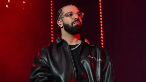 Drake pictured on stage in 2022. He wears an oversized leather jacket over a black top and diamond necklace. He has clear-framed square glasses and a trimmed beard. The staging behind him is lit red. 