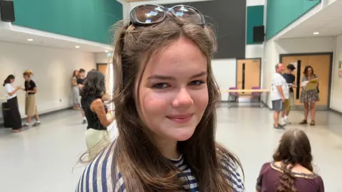 BBC/ Elizabeth Baines A young woman in a stripey top smiles from a school hall on results day.
