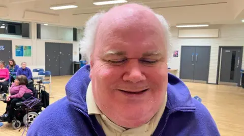 Gareth is smiling at the camera. He is wearing a blue fleece with a beige top underneath. He has white wispy hair around the side of his head. 