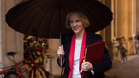 Getty Images Emma Reynolds, a white woman aged 48, walking to Downing Street. She is holding a large black umbrella. She is wearing a navy suit jacket with red trim and carrying a red folder. She has short blonde hair