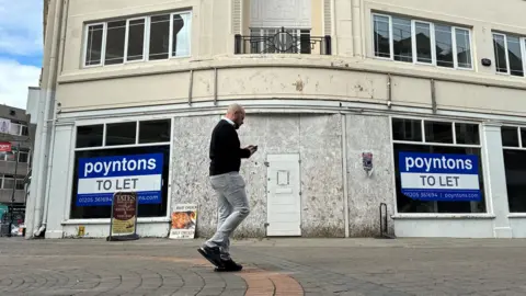 Sharon Edwards/BBC A man walks in front of an empty shop. The entrance is boarded up and  large 