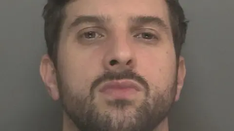 A mugshot of Thomas Cashman, who has dark brown hair and black stubble, and purses his lips at the camera with a defiant expression