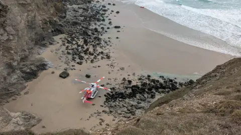 A red and white helicopter on a beach at the bottom of cliffs.