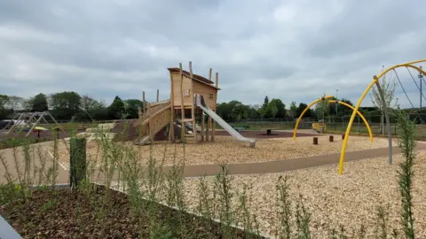 Reigate & Banstead Borough Council A wooden play area with a slope leading up to one side of a building on long legs and a metal slide coming out another side. The floor is woodchipped and there are yellow metal bar structures with swings on them.