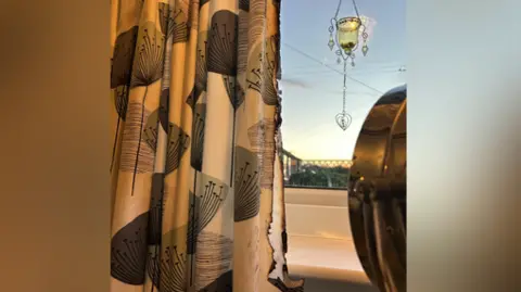 A fire service picture showing part of a magnifying glass in the foreground. It is pointing towards a set of curtains. The edges of the curtains are singed. Bright sky can be seen through the window.