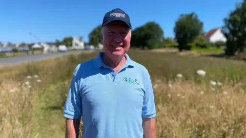 Gordon is standing in a field and smiling at the camera. He is wearing a navy blue baseball cap and a light blue polo t-shirt.