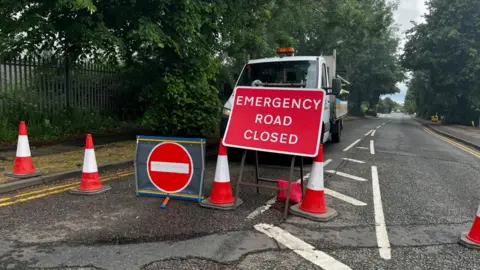 Emma Baugh/BBC A road closure, showing a red sign with white lettering "EMERGENCY ROAD CLOSED", a line of cones and a "no entry" sign. A triangular hashed area is adjacent to the line of cones on the road. A white lorry can be seen behind the closure. There are trees either side of the road and a wooden fence to the left.