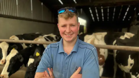 BBC Ryan Taggart standing with his arms crossed looking at the camera. In the background there are dairy cows eating in a shed. 