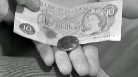 A black and white image of a hand holding a new 50p coin on top of an old ten-shilling note.