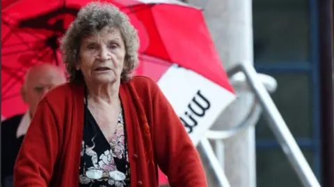 Sheila is wearing a red cardigan over a floral dress. She has grey/brown curly hair. 