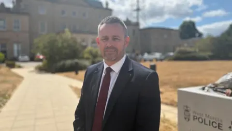 A man wearing a dark suit and a burgundy tie with dark grey hair, stands outside the West Mercia Police Head Quarters