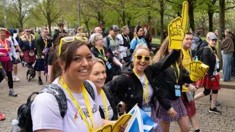 Elaine Livingstone Kiltwalkers wearing yellow sunglasses and holding large pointing yellow foam hand smiling at the camera. There are more walkers behind them.