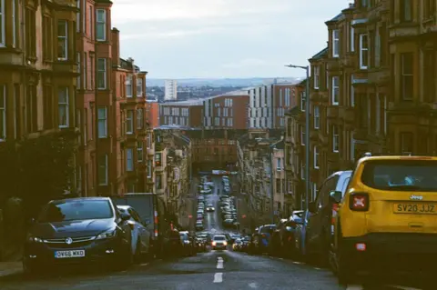 Rory Ballantyne A downhill residential street lined with multi‑storey tenement buildings, with parked cars on both sides and light traffic visible in the distance. 