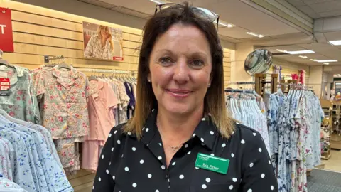 Diane smiles for the camera, taken in the shop. She has brown hair, with glasses pushed up over her head. She wears a polka dot top.