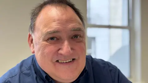 David Storm. He has short receding salt and pepper hair and blue eyes. He is wearing a blue shirt and smiling at the camera. He is in a room with a window behind him and the background is blurred.