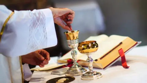 Getty Images An alter with a hand dipping communion bread into a goblet of wine, and an open bible open behind.