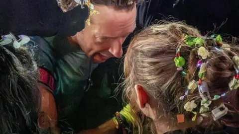 Isla Chris Martin is bending down close to Isla and is talking to her. Her face is turned away from the camera. The bun in her blonde hair is covered with flowers and beads.