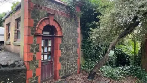 Malvern Hills District Council A stone-fronted toilet block with a brown door, framed by red bricks. There is ivy growing down one wall and ivy-covered trees in front of it.