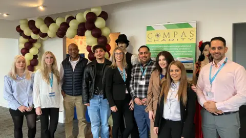 Shariqua Ahmed/BBC Petr Torak extreme right in a pink shirt and grey trousers standing in a group with his team in front of a yellow and green signboard 