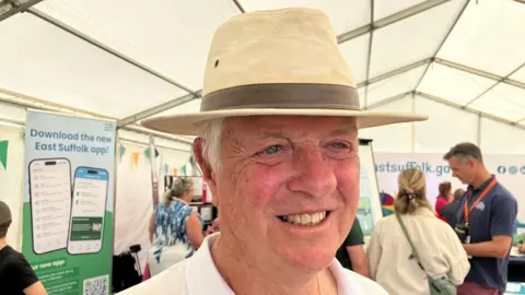 Vikki Irwin/BBC A man wearing a cream coloured hat with a large and wide brim smiles at the camera. Some grey hair can be seen underneath the hat. He is wearing a white shirt. He is standing inside a large white tent during an agricultural show. People are talking behind him. 