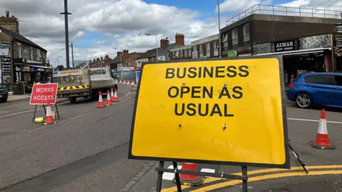 JOANNA TAYLOR/BBC A yellow road sign which reads 'Business Open as Usual' stands on a road. Behind it is a road with cones, a maintenance vehicle and more road signs.