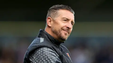 Getty Images Jon Brady wearing a black Northampton Town tracksuit. He has gelled dark hair and is looking into the distance with his head tilted sideways. 