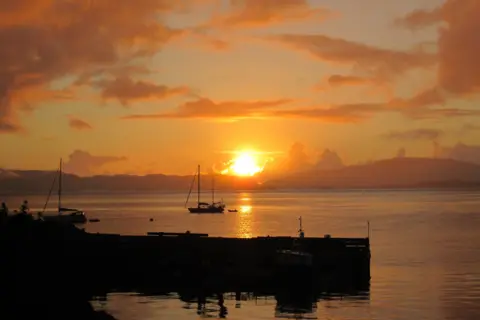 Richard Hadlow An orange sunset over a harbour, with sailing boats in the water.