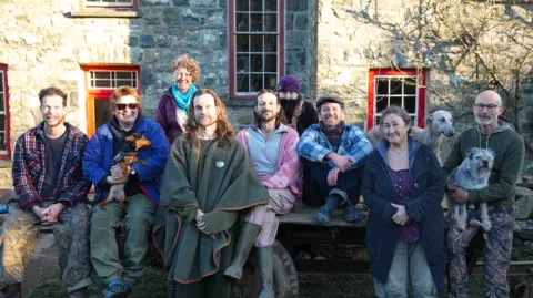 The community at Brithdir Mawr sitting in a group outside the large old stone house that has red leaded windows. There are nine people of mixed ages and three dogs.