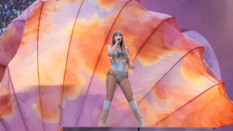 PA Media Taylor Swift on stage at a concert in London, in front of a large peach and a light purple background. She is singing into a microphone and wearing a one-piece and knee-high boots.