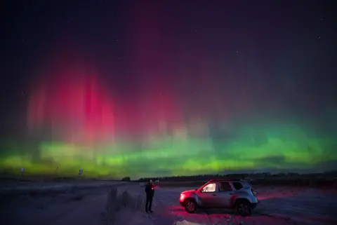 Alexander Manzyuk/Anadolu via Getty Images Seorang pria, memegang obor, berdiri di samping kendaraan - dengan lampu interior menyala - untuk mengamati Cahaya Utara, tampak berwarna merah muda dan hijau di langit di atas kota Krasnoyarsk di Siberia, Rusia pada 2 Januari , 2025. 