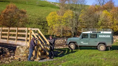 Yorkshire Dales National Park Authority Two men fix a wooden bridge over a stream behind are green trees and hills. A dark green rangers 4x4 is parked behind them with the word 'RANGER' written in white on the side