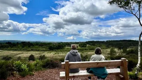Don Don Rear view of two people sitting on a wooden bench look out across a picturesque rural landscape under a blue sky dotted with white clouds