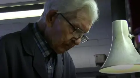 A man, Stan Shaw, with grey hair, wearing glasses and blue overalls in a workshop. He is looking down as if working at something on his bench. It is a close up showing only his head.