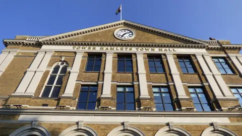 Tower Hamlets Town Hall taken from a low angle. A clock is positioned at the top of the building with a flag on top. 