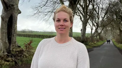 Elsie is smiling at the camera, she has blonde hair tied up. She is wearing a white jumper and the trees can be seen in the background. 