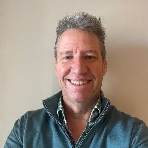 Andy Barton smiles at the camera - he is wearing a blue zipper top over a green and white check shirt and stands against a cream-coloured wall