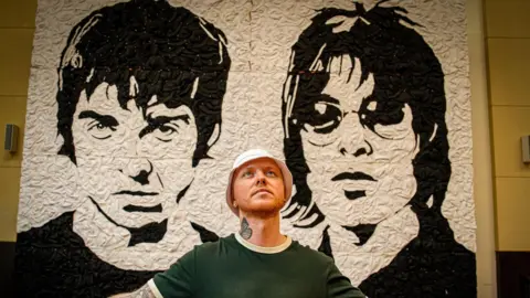 The artist standing with his arms outstretched at though on the cross with the full mural in the background. He is dressed the same, but has on a white bucket hat.