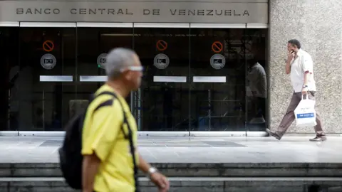 A person wearing a yellow T shirt and a backpack walks past the central bank of Venezuela as another person in a white shirt walks holding a shopping bag in the background, outside a glass entrance with the words 'Banco Central de Venezuela' on it, in Caracas in 2017.
