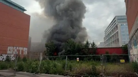 Smoke billowing from a fire, in a huge plume going upward, behind a wall which is behind a metal fence.