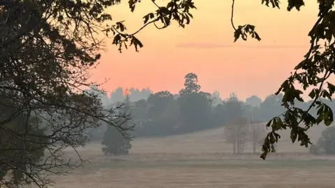 A photo of a rural field taken early in the morning, shows a grey haze of smoke in the air. The sun can just be seen rising behind trees in the distance, making the sky orange.
