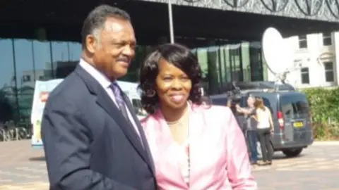 Dr. Beverly Lindsay A man in a suit with short dark hair alongside a woman in a pink jacket with dark hair in front of a large, glass-fronted building
