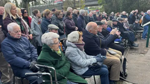 A large crowd of people outside. Some are sitting and behind them there are rows people standing.