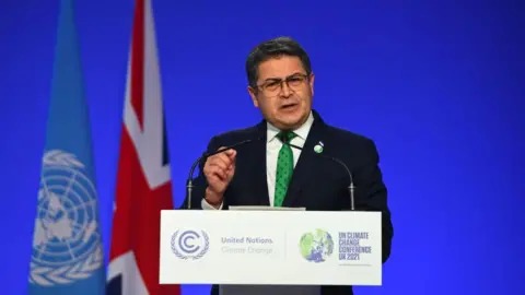 Getty Images Honduras President Juan Orlando Hernández speaking at a podium, wearing a black suit and a green dotted tie. Behind him is a blue backdrop with a UN flag and a UK flag to his left. He has short hair and wears square-framed eye glasses. 