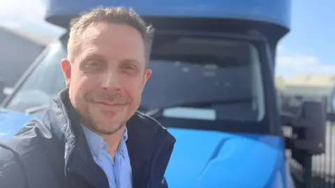 A man with short, grey hair smiles and takes a selfie. He has a blue shirt and a dark coat on. He stands in front of a blue van.